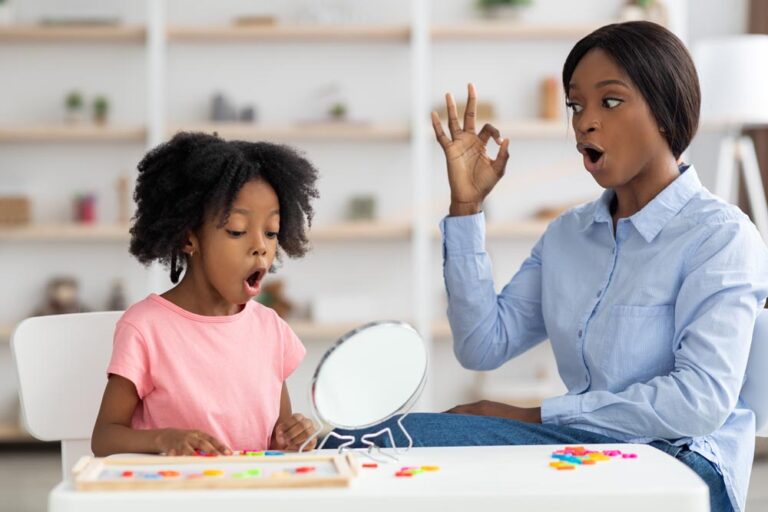 Jolie petite fille noire assistant à une séance d'orthophonie à la clinique, se regardant dans un miroir, belle jeune femme afro-américaine orthophoniste travaillant sur les sons avec l'enfant pour lui enseigner la prononciation de ch et sch en allemand.